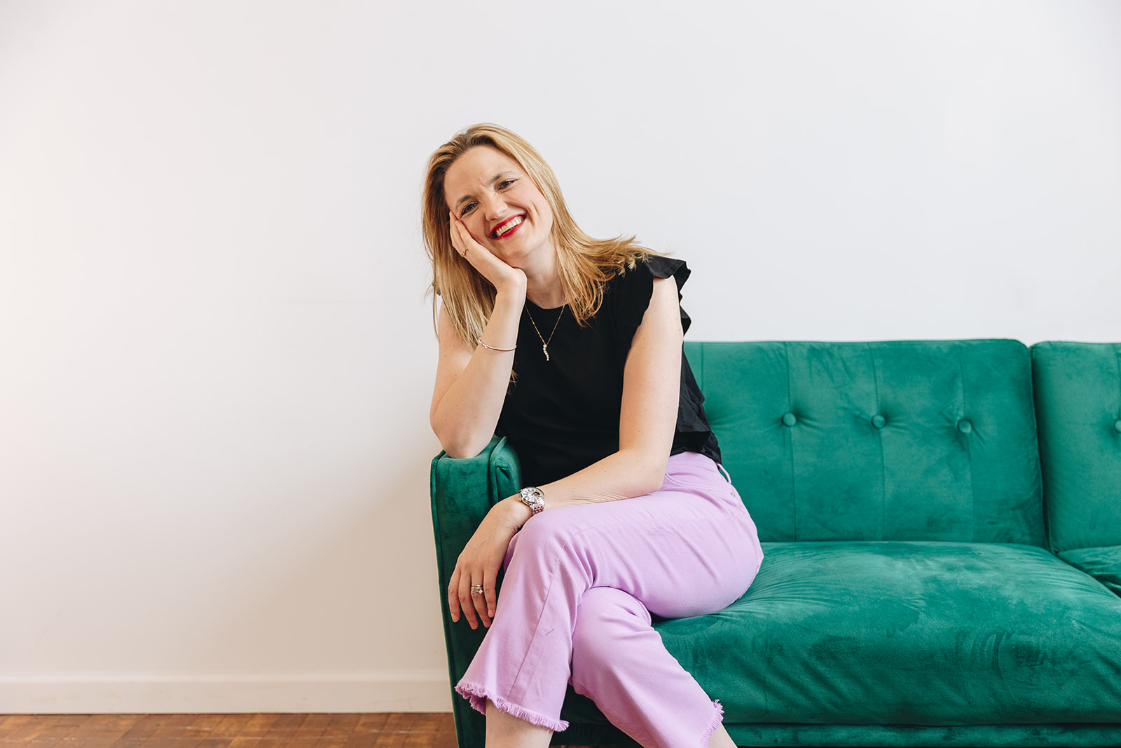 Brooke Greening, a sales coach, sitting on a green couch, smiling, and wearing a black top and purple pants.