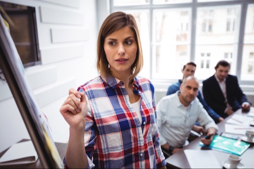 Woman in a plaid shirt giving a presentation to a group of colleagues in a conference room.